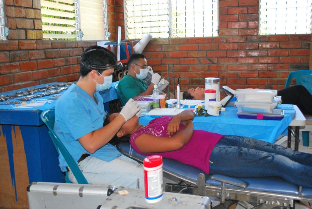 image description: two men in nursing scrubs and surgical masks perform dental care tasks on two young people in a rustic brick-walled clinic. 