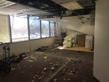 image description: A large suburban office room is in a state of disarray mid-demolition. Portions of the ceiling are missing and debris is scattered across the floor. Sunlight streams in through a wall of windows. 