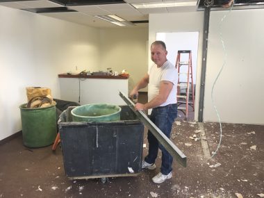 image description: A man in jeans and a white t-shirt stands in a partially demolished office, working on a steel beam with a small hand tool. 