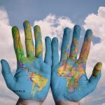 A pair of hands with a world map painted on the palms, held up against a blue sky with puffy white clouds
