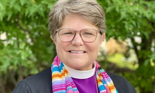 A headshot of Bishop DeDe Duncan-Probe+. Bishop DeDe is outside in front of a leafed-out, lusciously green tree in mid-summer. She's smiling at the camera, wearing her glasses, her collar and Bishop-purple shirt, a multi-colored scarf, and her pectoral cross.
