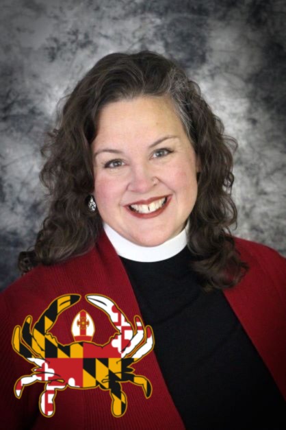 A headshot of Bishop-Elect Carrie Schofield-Broadbent smiling to camera. She's wearing her clerical collar, a black shirt, and a deep red cardigan. Super imposed in the bottom left corner of the image is a rather clever logo from the Diocese of Maryland for their Bishop Search. A crab, colored with the Maryland flag and its bold gold/black and red/white patterns, is wearing a mitre! 