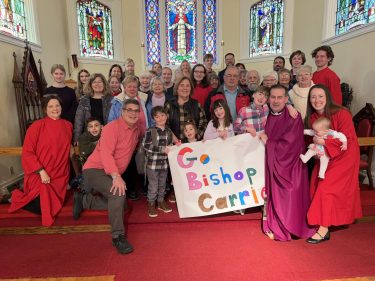 A group of smiling parishioners of all ages are standing in the sanctuary at St. John's Marcellus, Bishop-Elect Carrie's home church, and holding a beautiful, colorful sign that reads "Go Bishop Carrie!" 