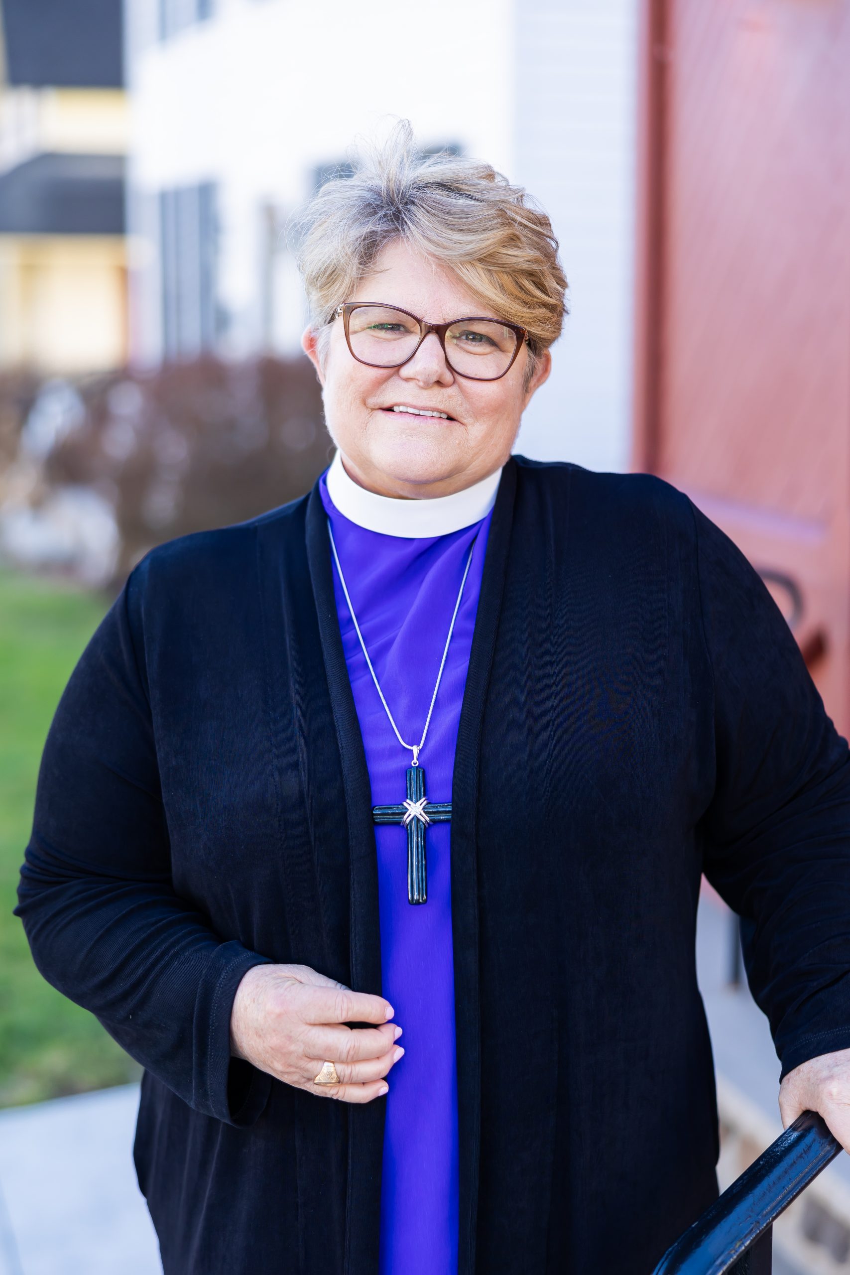 Bishop DeDe smiles broadly and kindly toward the camera. She's wearing her clericals, including her collar and a pectoral cross. She's outdoors on the steps of a church. You can see a hint of the recognizably Episcopal-Red door over her left shoulder.