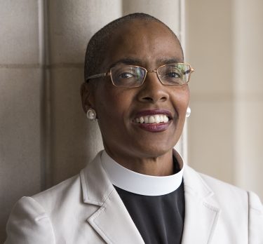 Image shows the Rev. Kelly Brown Douglas, Ph.D smiling. She is wearing a white collar with black shirt and an off-white blazer leaning against a while marble wall and column