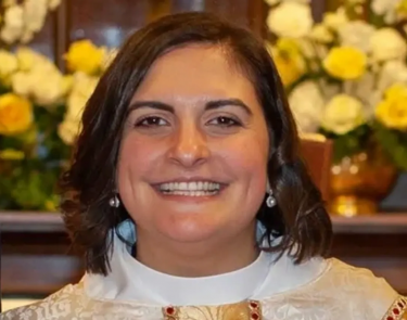 Image Description: A photo of Rev. Pilar. A woman with shoulder-length dark hair smiles warmly at the camera. She is wearing clerical attire, including a white robe with ornate patterns. The background is adorned with yellow and white flowers, adding to the serene and cheerful atmosphere.